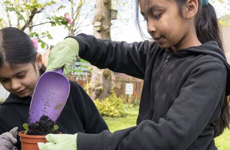 two pupils wearing black pour soil into a plant pot wearing green gardening gloves outside near a tree