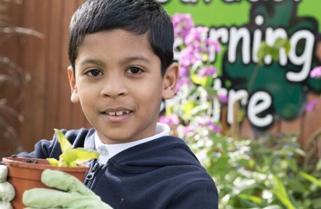 a pupil holding a plant pot with green gloves with outdoor learning centre sign behind